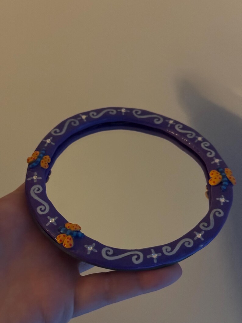 May include: A round, purple mirror with a decorative border. The border features white swirls, crosses, and small, colorful butterfly accents. The mirror is held in a hand.