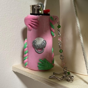 Op de afbeelding: Een roze aansteker met groene en roze klei versieringen, waaronder handen en gedraaide touwontwerpen. Een zilverkleurige bedelketting met groene en roze kralen en een stervormige bedel hangt aan de aansteker.