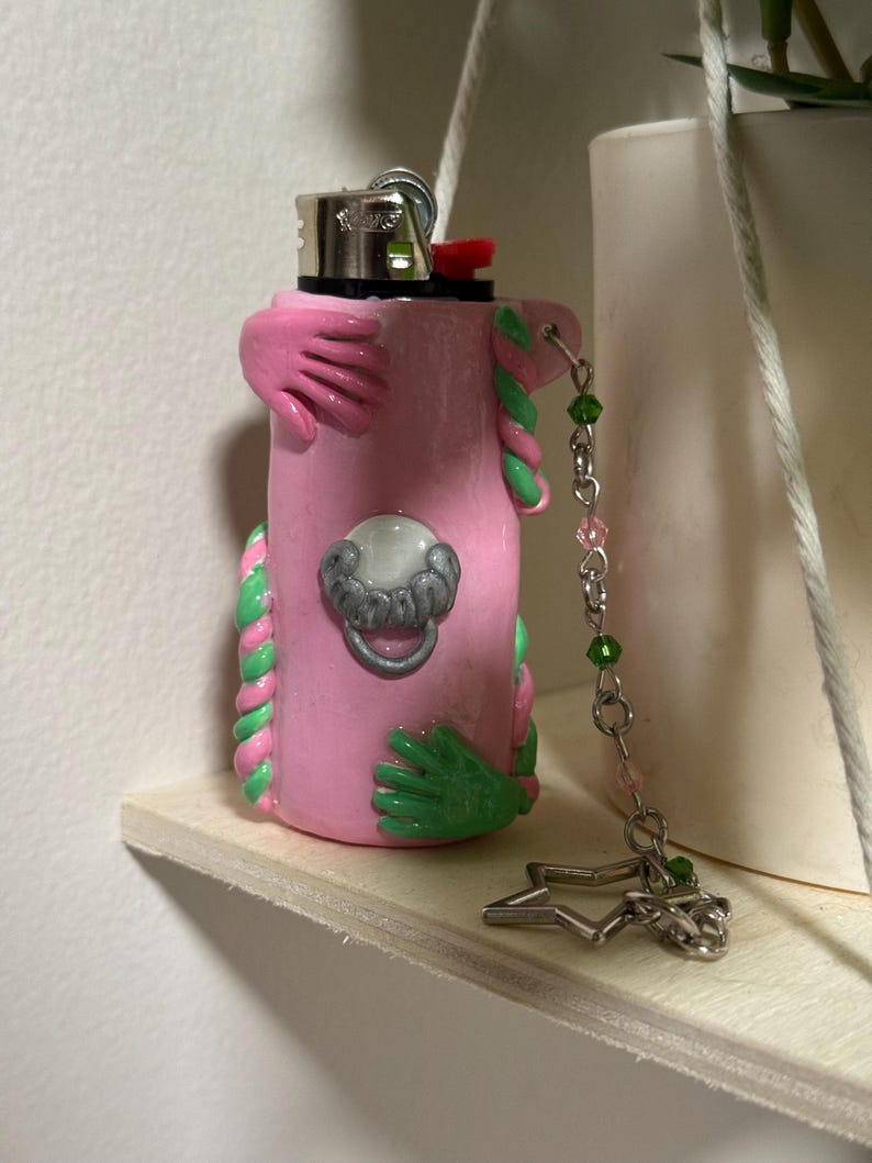 Op de afbeelding: Een roze aansteker met groene en roze klei versieringen, waaronder handen en gedraaide touwontwerpen. Een zilverkleurige bedelketting met groene en roze kralen en een stervormige bedel hangt aan de aansteker.