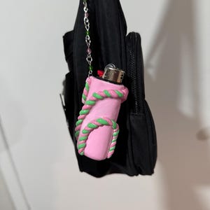 Op de afbeelding: Een roze aansteker met groene en roze touwachtige details. De aansteker is bevestigd aan een zwarte tas met een kralenketting. De aansteker is een uniek accessoire.