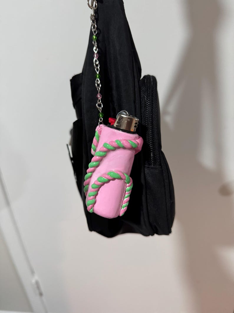 Op de afbeelding: Een roze aansteker met groene en roze touwachtige details. De aansteker is bevestigd aan een zwarte tas met een kralenketting. De aansteker is een uniek accessoire.