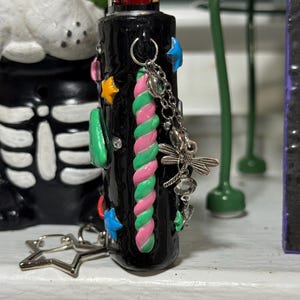 May include: Black lighter decorated with colorful embellishments. Features a pink, green, and white twisted design, along with star and blue star-shaped accents. A silver charm with a dragonfly and other charms hangs from the lighter.