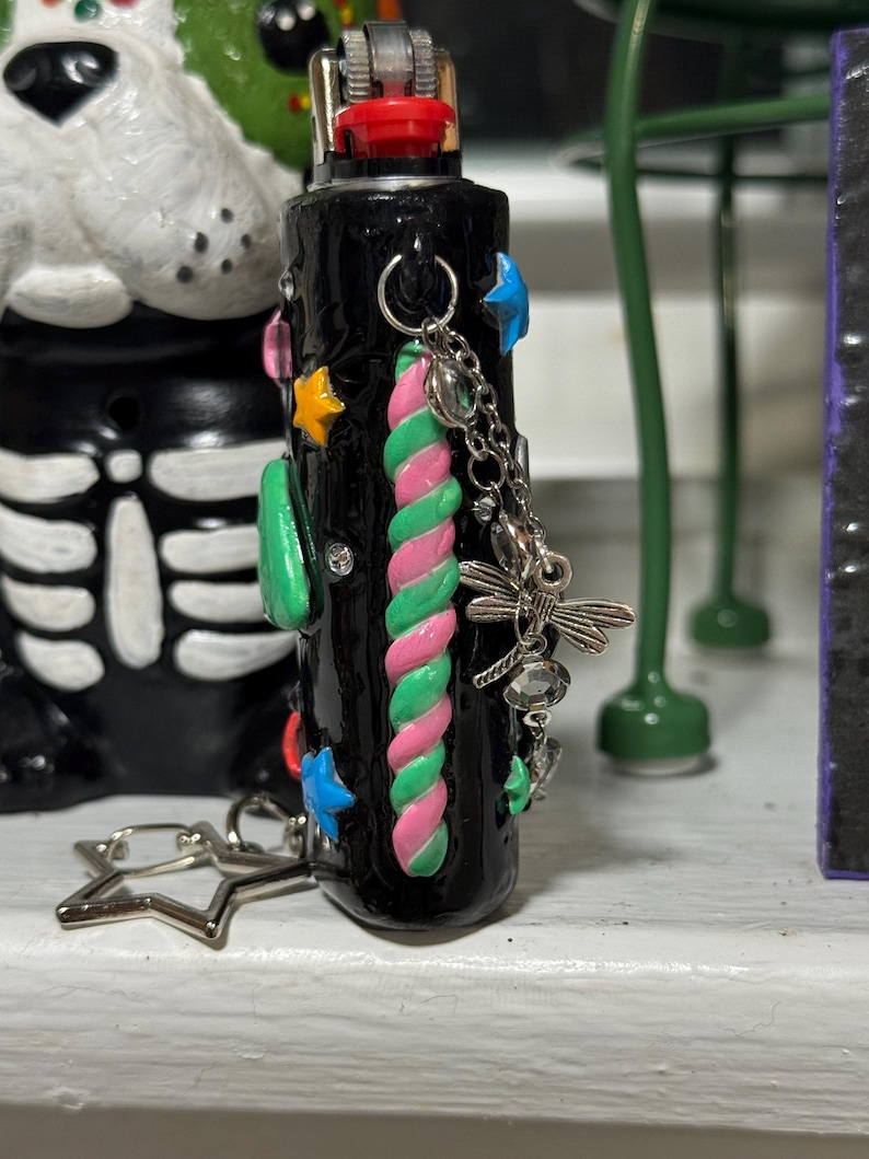 May include: Black lighter decorated with colorful embellishments. Features a pink, green, and white twisted design, along with star and blue star-shaped accents. A silver charm with a dragonfly and other charms hangs from the lighter.
