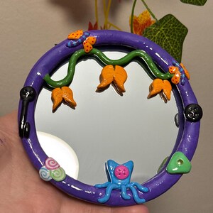 May include: A round, purple-framed mirror with whimsical clay embellishments. The frame features a green vine with orange flower-like shapes, butterflies, and a blue octopus-like creature. Additional details include a black key, buttons, and a green triangle.