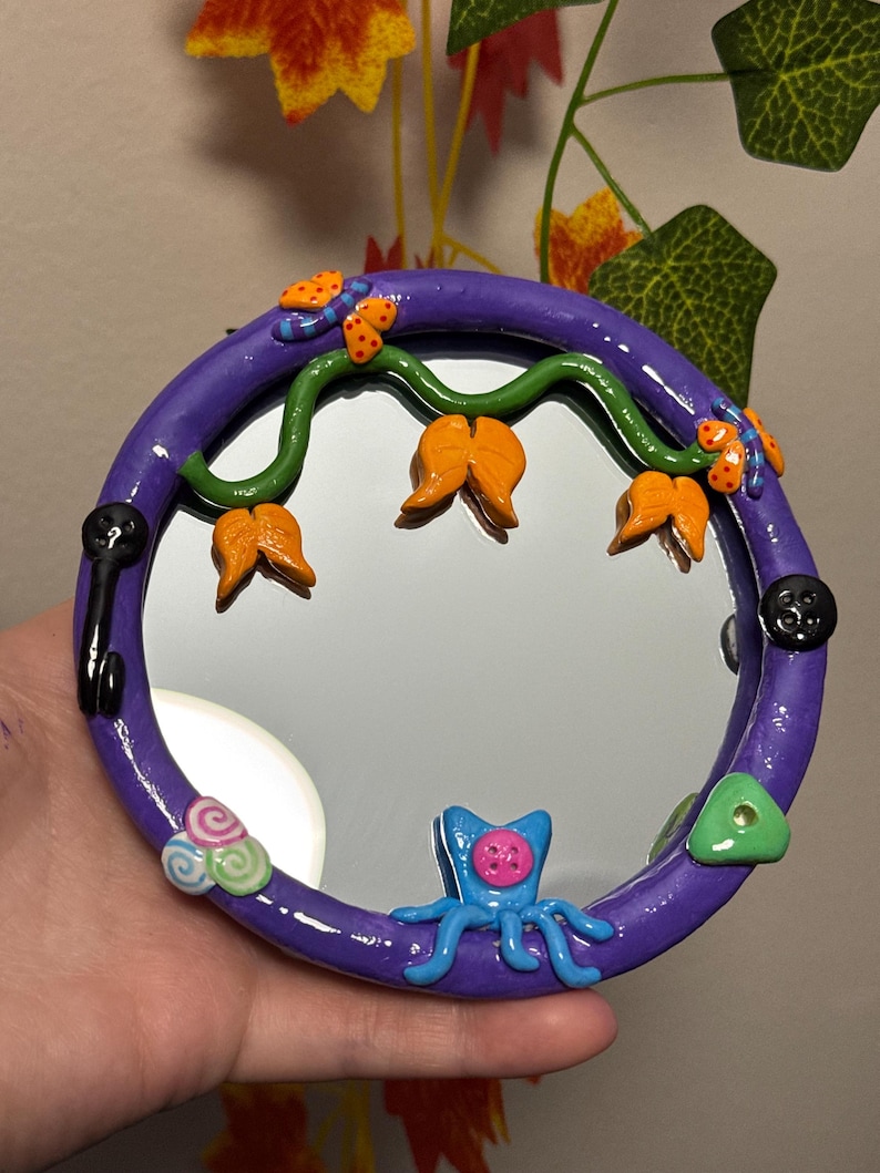 May include: A round, purple-framed mirror with whimsical clay embellishments. The frame features a green vine with orange flower-like shapes, butterflies, and a blue octopus-like creature. Additional details include a black key, buttons, and a green triangle.