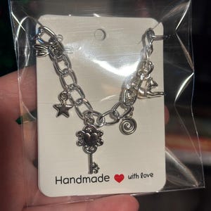 May include: Silver charm bracelet with a key, star, and spiral charms. The bracelet is presented on a white card with the text "Handmade with love" and a red heart. The bracelet is in a clear plastic bag.