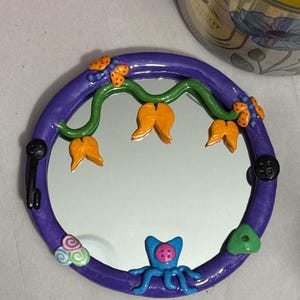 May include: A round, purple-framed mirror with whimsical clay embellishments. The frame is decorated with orange flower-like shapes, green vines, and a blue octopus-like creature. Black button-shaped accents and colorful swirls add to the playful design.