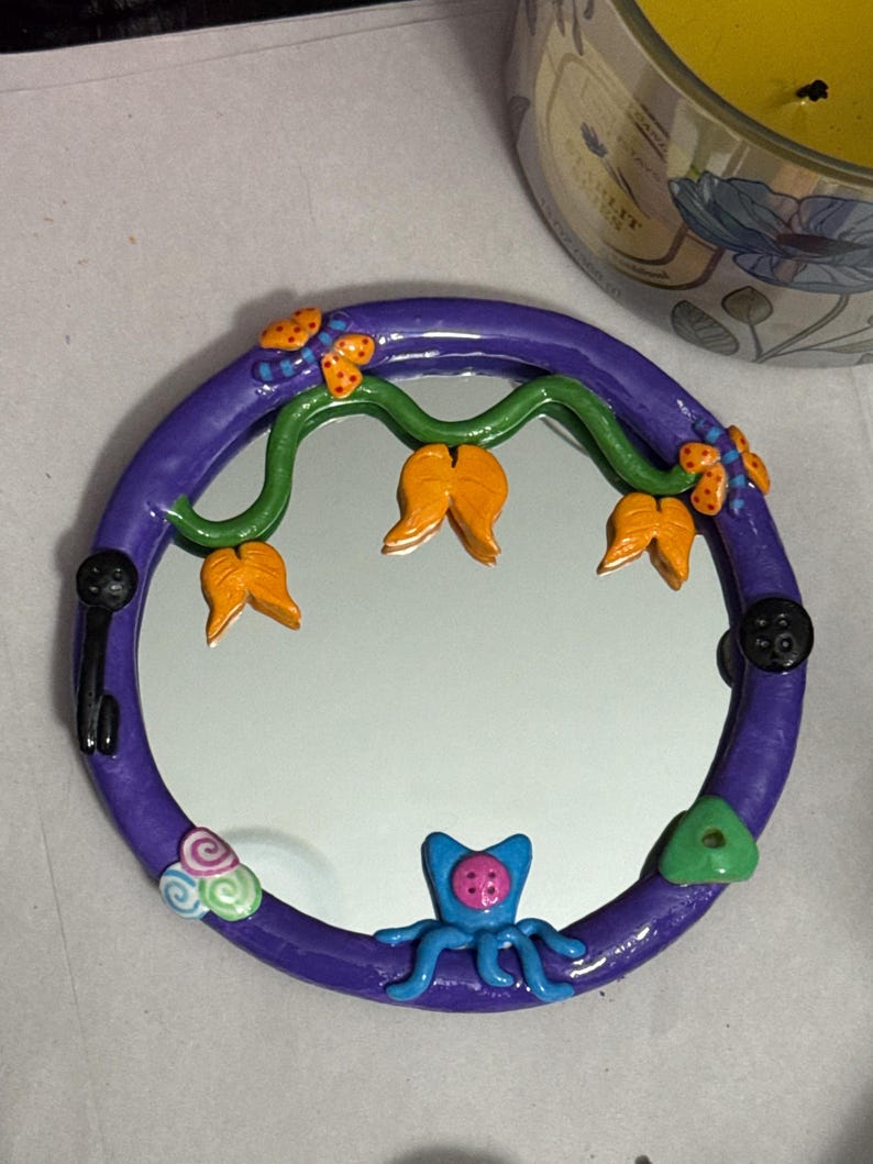 May include: A round, purple-framed mirror with whimsical clay embellishments. The frame is decorated with orange flower-like shapes, green vines, and a blue octopus-like creature. Black button-shaped accents and colorful swirls add to the playful design.