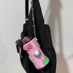 Op de afbeelding: Een roze aanstekerhoesje met groene en roze accenten, met een ontwerp van handen en een zilveren embleem. De aansteker is bevestigd aan een zwarte tas met een ketting van roze en groene kralen.
