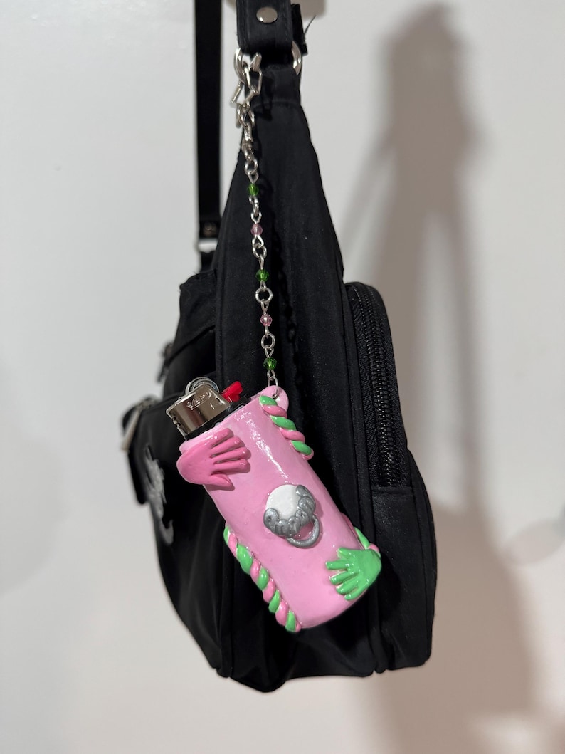 Op de afbeelding: Een roze aanstekerhoesje met groene en roze accenten, met een ontwerp van handen en een zilveren embleem. De aansteker is bevestigd aan een zwarte tas met een ketting van roze en groene kralen.