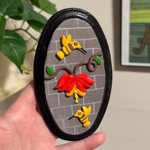May include: Oval-shaped wall art featuring a brick background and a black frame. The artwork depicts a red and yellow flower with two yellow hummingbirds. The hummingbirds have black beaks and are positioned around the flower.