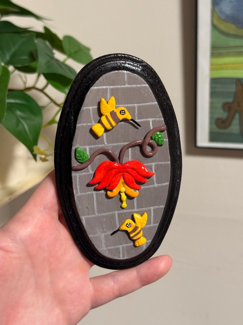 May include: Oval-shaped wall art featuring a brick background and a black frame. The artwork depicts a red and yellow flower with two yellow hummingbirds. The hummingbirds have black beaks and are positioned around the flower.