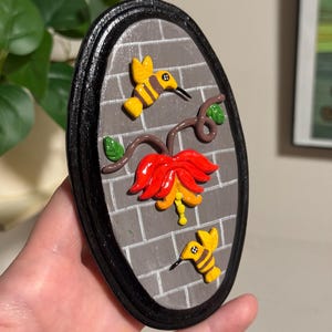 May include: Oval-shaped wall art featuring a red and yellow flower with two yellow hummingbirds and green leaves. The background is a gray brick pattern, framed by a black border.