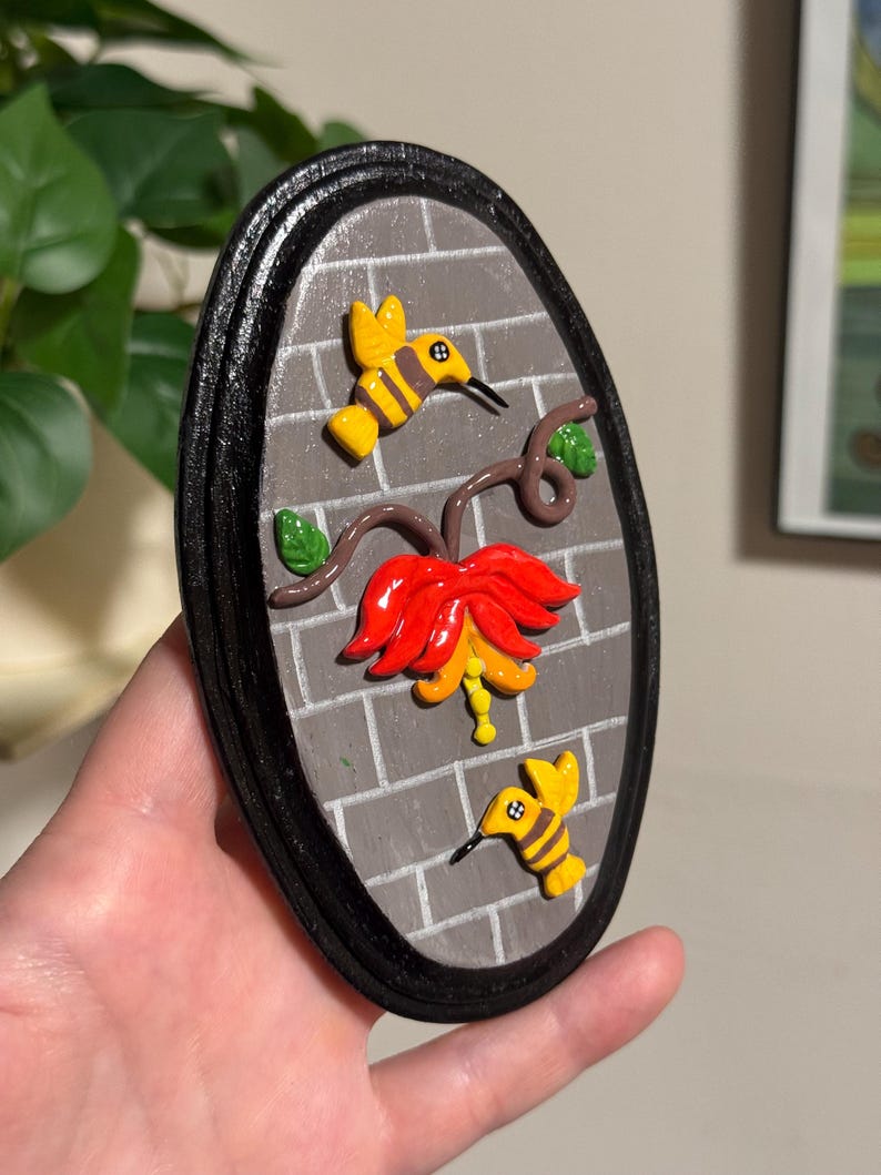 May include: Oval-shaped wall art featuring a red and yellow flower with two yellow hummingbirds and green leaves. The background is a gray brick pattern, framed by a black border.