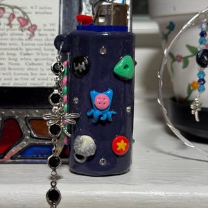 May include: A navy blue lighter decorated with colorful, whimsical embellishments. The lighter features a red button, a blue top, and various attached charms, including a dragonfly and black beads. The lighter is adorned with small, colorful shapes.