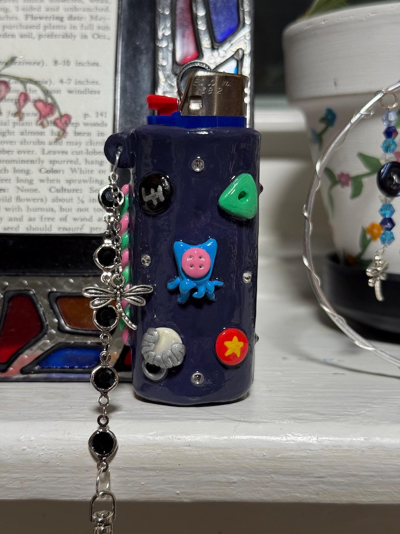 May include: A navy blue lighter decorated with colorful, whimsical embellishments. The lighter features a red button, a blue top, and various attached charms, including a dragonfly and black beads. The lighter is adorned with small, colorful shapes.