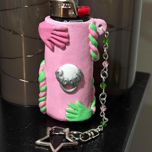 Op de afbeelding: Een roze aansteker met groene en roze accenten, met gebeeldhouwde handen en een decoratieve zilveren bedel. De aansteker heeft een zilveren ster-vormige bedel bevestigd aan een ketting met groene en roze kralen. De aansteker is versierd met een zilveren sluiting.