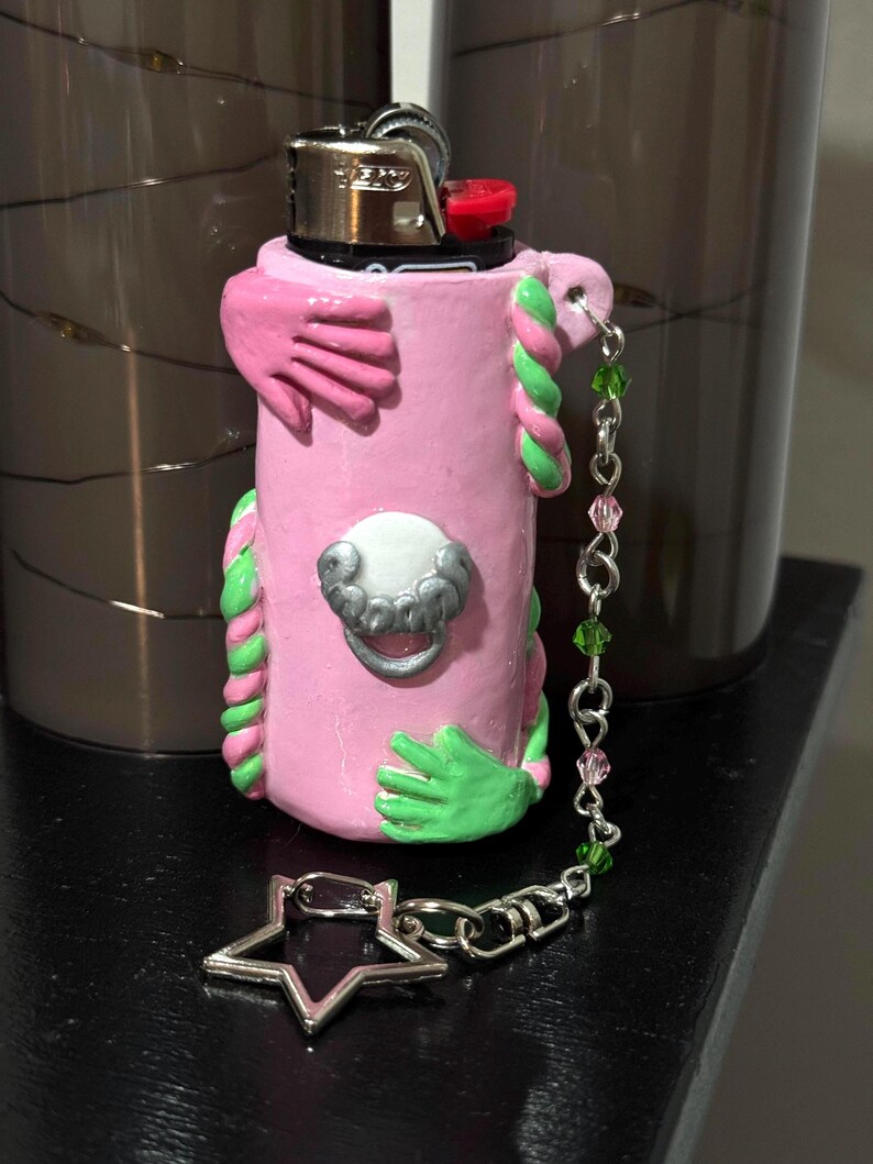 Op de afbeelding: Een roze aansteker met groene en roze accenten, met gebeeldhouwde handen en een decoratieve zilveren bedel. De aansteker heeft een zilveren ster-vormige bedel bevestigd aan een ketting met groene en roze kralen. De aansteker is versierd met een zilveren sluiting.