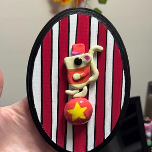 Puede incluir: Decoraci&oacute;n de pared ovalada con un rat&oacute;n de circo equilibr&aacute;ndose sobre una bola roja con una estrella amarilla. El rat&oacute;n lleva un sombrero y una chaqueta rojos. El fondo tiene rayas rojas y blancas, enmarcado por un borde negro.