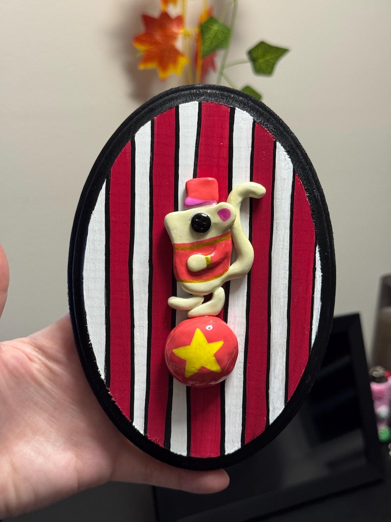 Puede incluir: Decoraci&oacute;n de pared ovalada con un rat&oacute;n de circo equilibr&aacute;ndose sobre una bola roja con una estrella amarilla. El rat&oacute;n lleva un sombrero y una chaqueta rojos. El fondo tiene rayas rojas y blancas, enmarcado por un borde negro.