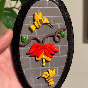May include: Oval-shaped wall art featuring a gray brick background with a black border. Two yellow and black hummingbirds are depicted, along with a red and yellow flower and brown vines. Green leaves add detail to the design.