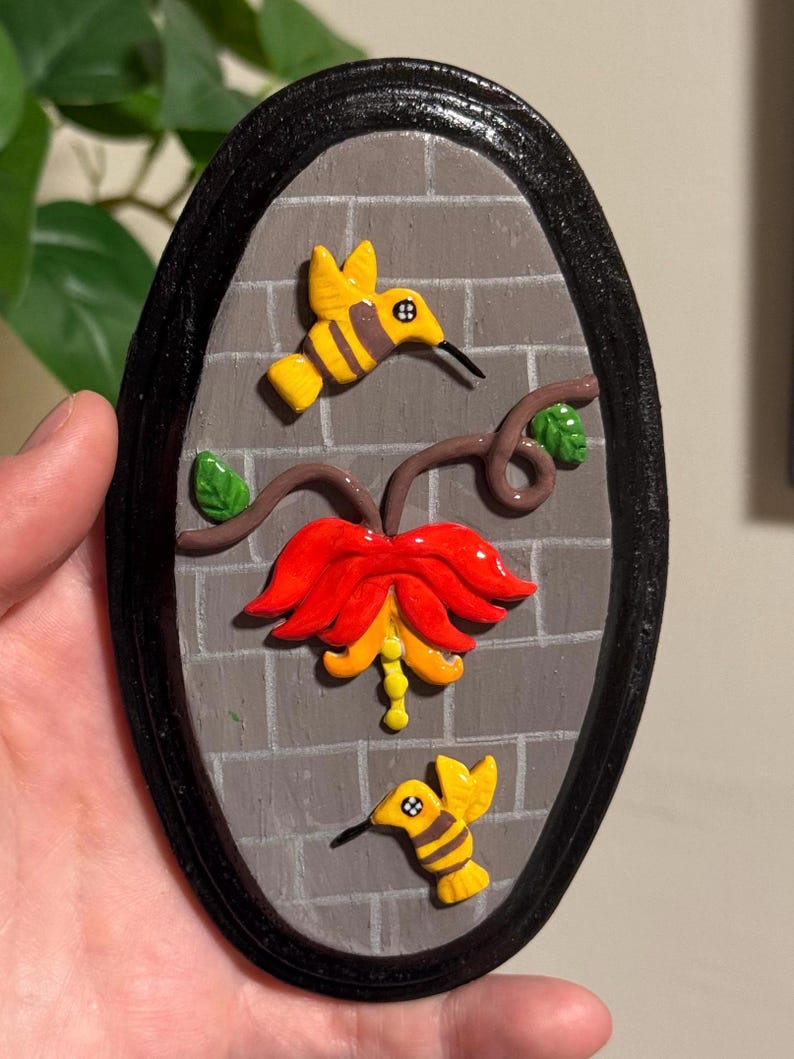 May include: Oval-shaped wall art featuring a gray brick background with a black border. Two yellow and black hummingbirds are depicted, along with a red and yellow flower and brown vines. Green leaves add detail to the design.