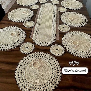 May include: A collection of cream-colored crochet table linens, including placemats, coasters, and a table runner, arranged on a dark wooden table. The placemats and runner have a scalloped edge design. The text "Maréa Croché" is visible.