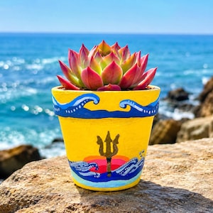 May include: A succulent plant with pink and green leaves potted in a yellow ceramic planter. The planter features a hand-painted design of blue ocean waves, a golden trident, and a sunset motif with orange and pink hues on the front surface.