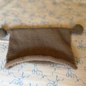 May include: A knitted brown hat with two small, round pom-poms on the ends. The hat is rectangular with a folded brim. The hat is made of a soft, textured yarn and is likely handmade. The hat is laying on a patterned surface.