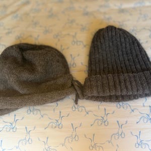 May include: Two knitted hats. One is a dark gray cloche style with a folded brim. The other is a ribbed knit beanie in a similar dark gray color. Both hats are made of a soft, textured yarn.