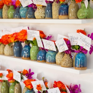 Puede incluir: Una exhibición de pequeños jarrones de cerámica coloridos, cada uno con flores vibrantes y una tarjeta con el nombre. Los jarrones son en tonos de azul, verde y beige moteado, dispuestos en estantes blancos. Las flores incluyen rosas naranjas y orquídeas moradas.