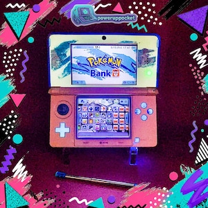 May include: A coral-colored Nintendo 3DS handheld game console, displaying the "Pokemon Bank" app on the top screen and a grid of app icons on the bottom screen. A stylus rests below the console. The background features a retro 80s and 90s design.