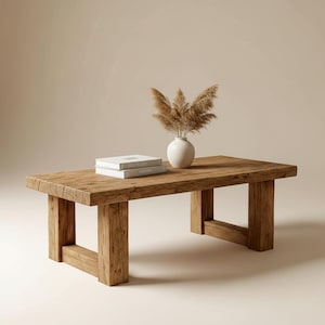 May include: A rectangular, rustic wooden coffee table with a natural finish. Two books and a small white vase with dried ornamental grass sit on the table. The table is supported by four thick, square legs.