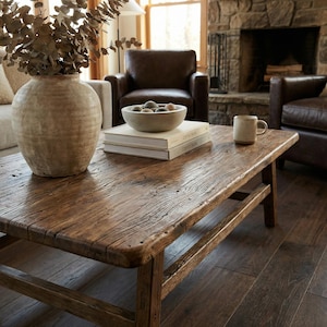 May include: A rustic, rectangular wooden coffee table with visible wood grain and a distressed finish. The table holds a large beige vase with dried branches, books, a bowl of decorative spheres, and a ceramic mug. The room features leather armchairs and a stone fireplace.