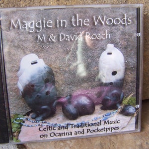 以下が含まれることがあります： M & David Roachによるアルバム「Maggie in the Woods」のCDケース。カバーアートは、石の背景の前に3つのオカリナを表示しています。「Celtic and Traditional Music on Ocarina and Pocketpipes」というテキストも表示されています。