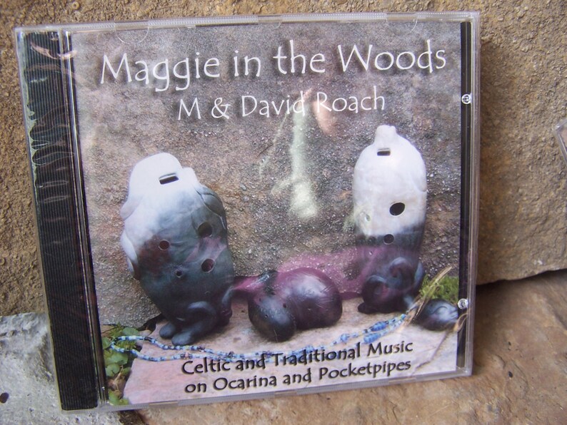以下が含まれることがあります： M & David Roachによるアルバム「Maggie in the Woods」のCDケース。カバーアートは、石の背景の前に3つのオカリナを表示しています。「Celtic and Traditional Music on Ocarina and Pocketpipes」というテキストも表示されています。
