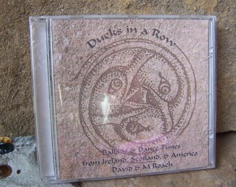 以下が含まれることがあります： ベージュの背景と茶色の円形のデザインが施されたCDケース。"Ducks in a Row"というテキストが黒色のフォントで書かれています。"Ballads & Dance Tunes from Ireland, Scotland, & America"というテキストが、より小さな黒色のフォントで書かれています。"David & M Roach"というテキストが、より小さな黒色のフォントで書かれています。