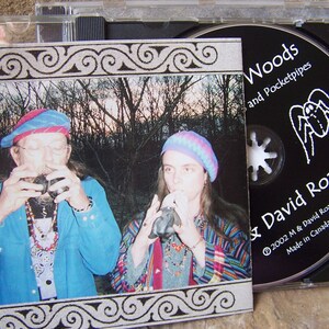 以下が含まれることがあります： 森の中で楽器を演奏する2人の写真が写ったCDケース。CDラベルにはM & David Roachの「Woods and Pocketpipes」と書かれています。アルバムのカバーには同じ画像が使われており、装飾的なボーダーが付いています。