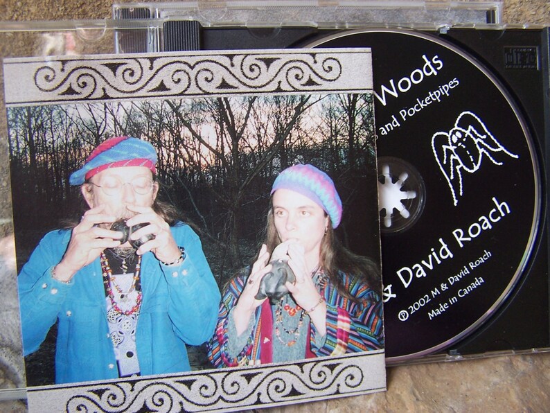 以下が含まれることがあります： 森の中で楽器を演奏する2人の写真が写ったCDケース。CDラベルにはM & David Roachの「Woods and Pocketpipes」と書かれています。アルバムのカバーには同じ画像が使われており、装飾的なボーダーが付いています。