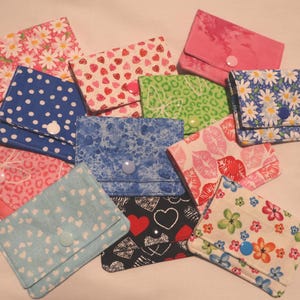 May include: A collection of small, handmade fabric wallets in various patterns and colors. Designs include daisies, hearts, polka dots, and abstract prints. Each wallet has a button closure and a unique fabric design.