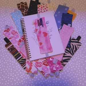 May include: A spiral-bound notebook with a pink pen holder and several fabric bookmarks. The bookmarks feature various patterns, including polka dots, hearts, and animal prints. The notebook has floral illustrations.