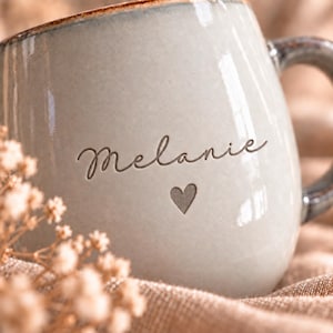 May include: A light gray ceramic mug with a glossy finish and a dark gray handle. The front of the mug features the name Melanie in a dark cursive script, positioned above a small, simple heart icon engraved into the surface of the ceramic material.