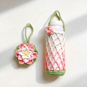 Könnte beinhalten: Ein gehäkelter Wasserflaschenhalter und ein passender Untersetzer. Der Flaschenhalter ist weiß, rosa und grün, mit einem floralen Akzent. Der Untersetzer ist eine rosa und weiße Blume mit grünem Boden und Schlaufe.