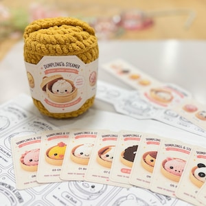 以下が含まれることがあります： 表面に質感のあるマスタードイエローの編み物製容器。Dumpling & Steamerと書かれた紙の帯が巻かれており、竹製の蒸し器に入った笑顔の点心のイラストが描かれています。長方形のコレクションカードが複数並び、様々な点心のキャラクターが描かれています。