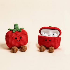 Two red crochet tomato-shaped cases, one closed and one open, with a green stem and smiling face. The open case contains white earbuds. Each case has two brown, ball-shaped feet. A whimsical design.