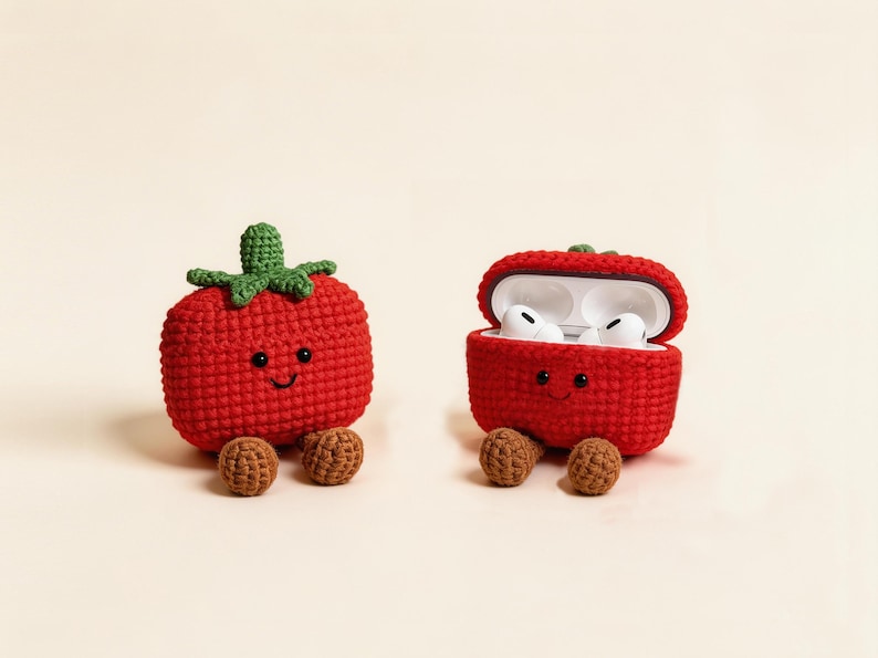 Two red crochet tomato-shaped cases, one closed and one open, with a green stem and smiling face. The open case contains white earbuds. Each case has two brown, ball-shaped feet. A whimsical design.