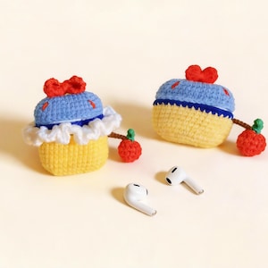 Two handmade crochet AirPod cases in a cupcake design. The cases are yellow with blue tops, white trim, and red bows. Each case has a red apple charm attached. Two white AirPods are also visible.