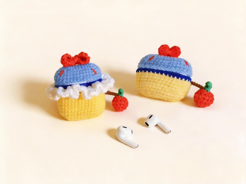 Two handmade crochet AirPod cases in a cupcake design. The cases are yellow with blue tops, white trim, and red bows. Each case has a red apple charm attached. Two white AirPods are also visible.