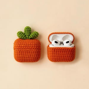 Two orange crochet cases, one closed and one open, designed to hold wireless earbuds. The closed case has a green crochet carrot top. The open case displays white earbuds. The cases are on a cream background.