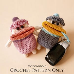 May include: Two crocheted duck toys, one pink and one grey, with large orange beaks and black eyes. The grey duck holds a car key. The text "CROCHET PATTERN ONLY" is at the bottom.